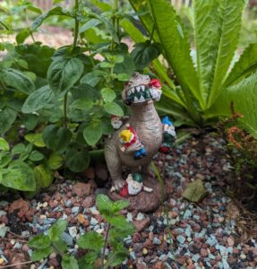 A garden ornament of a dinosaur eating gnomes. It is surrounded by plants.